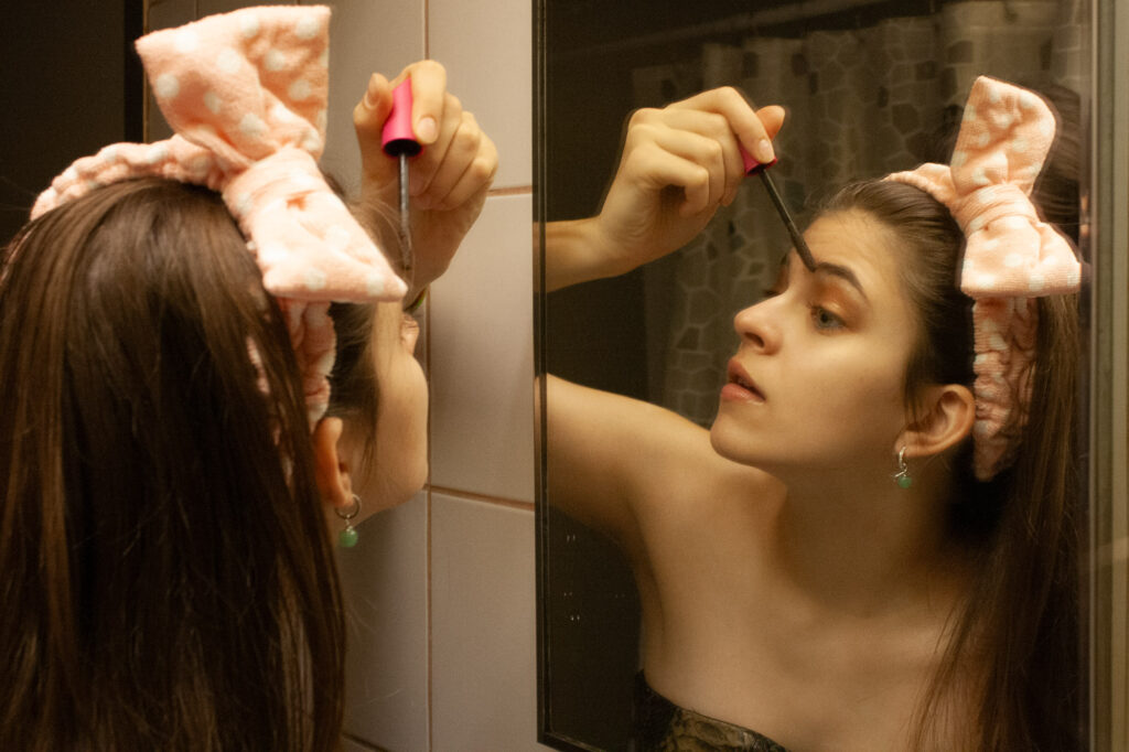 A woman doing her makeup in the washroom mirror.