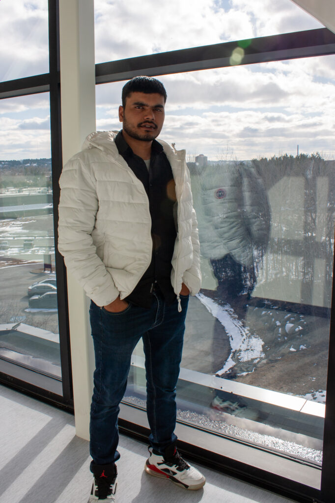 Man wearing a white coat, black shirt, and jeans posing in front of a bright window.